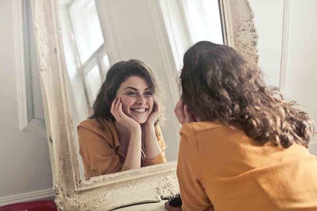 Девушка в светлой комнате смотрит на себя в зеркало и улыбается